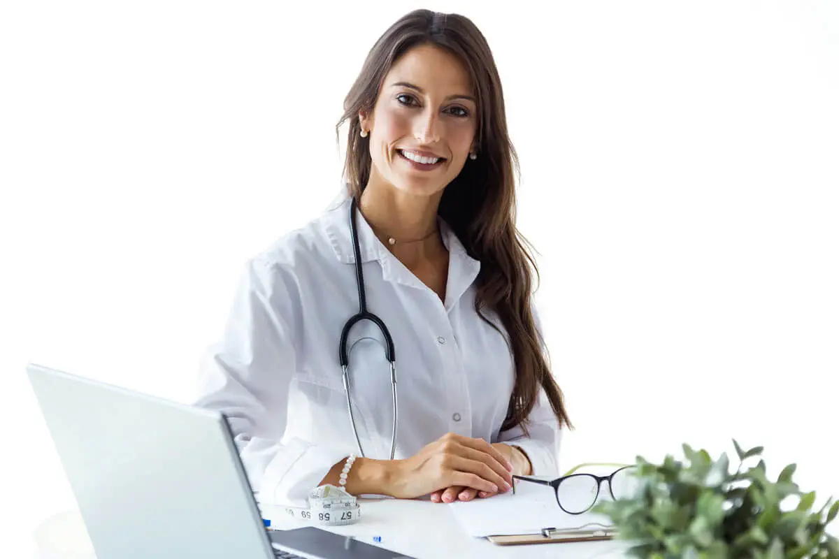 A smiling female doctor seated at a desk, wearing a white coat and stethoscope. A laptop, glasses, and a small plant are visible on the desk.